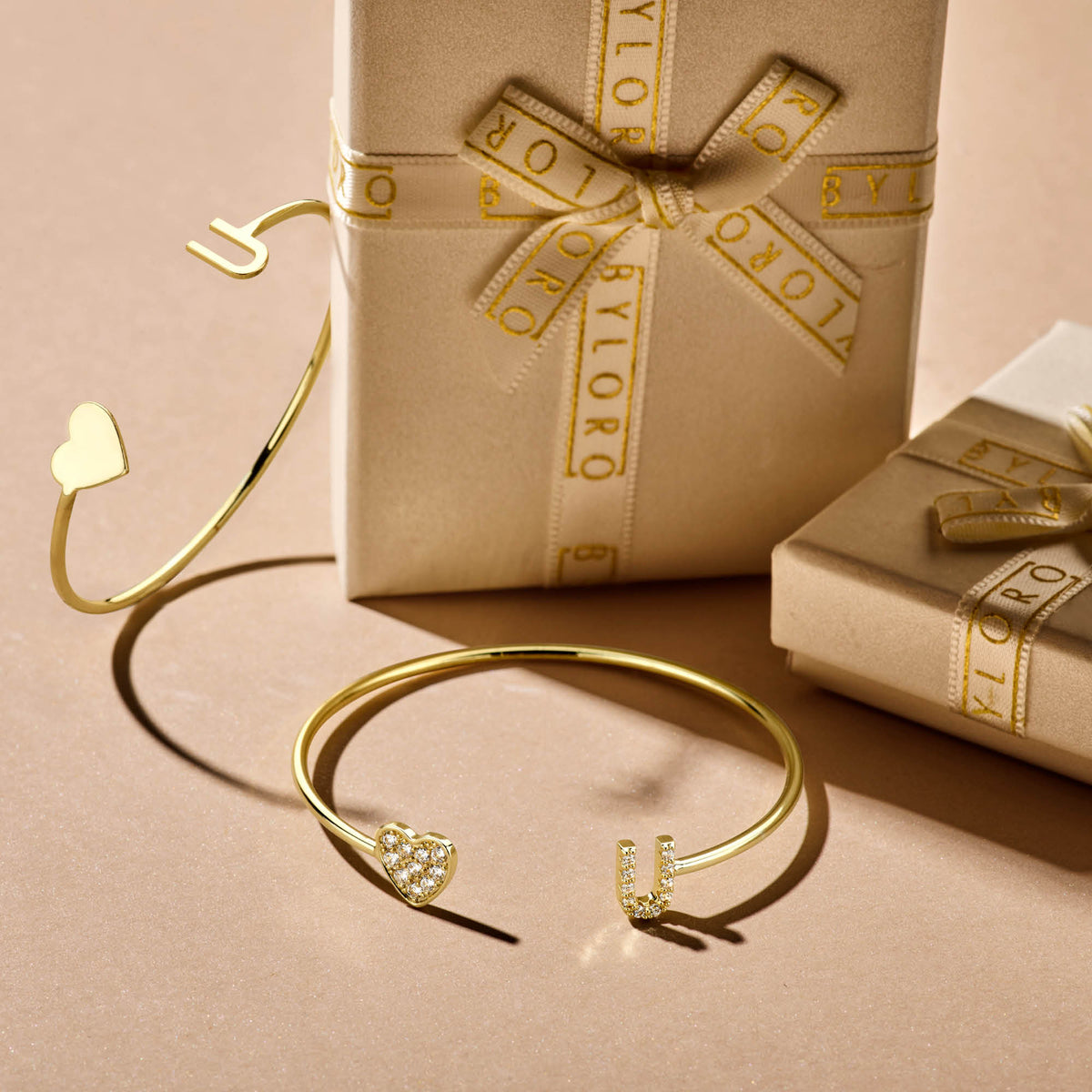 Twee goudkleurige open armbanden met hartjes en letters liggen naast beige Byloro-doosjes met lint.