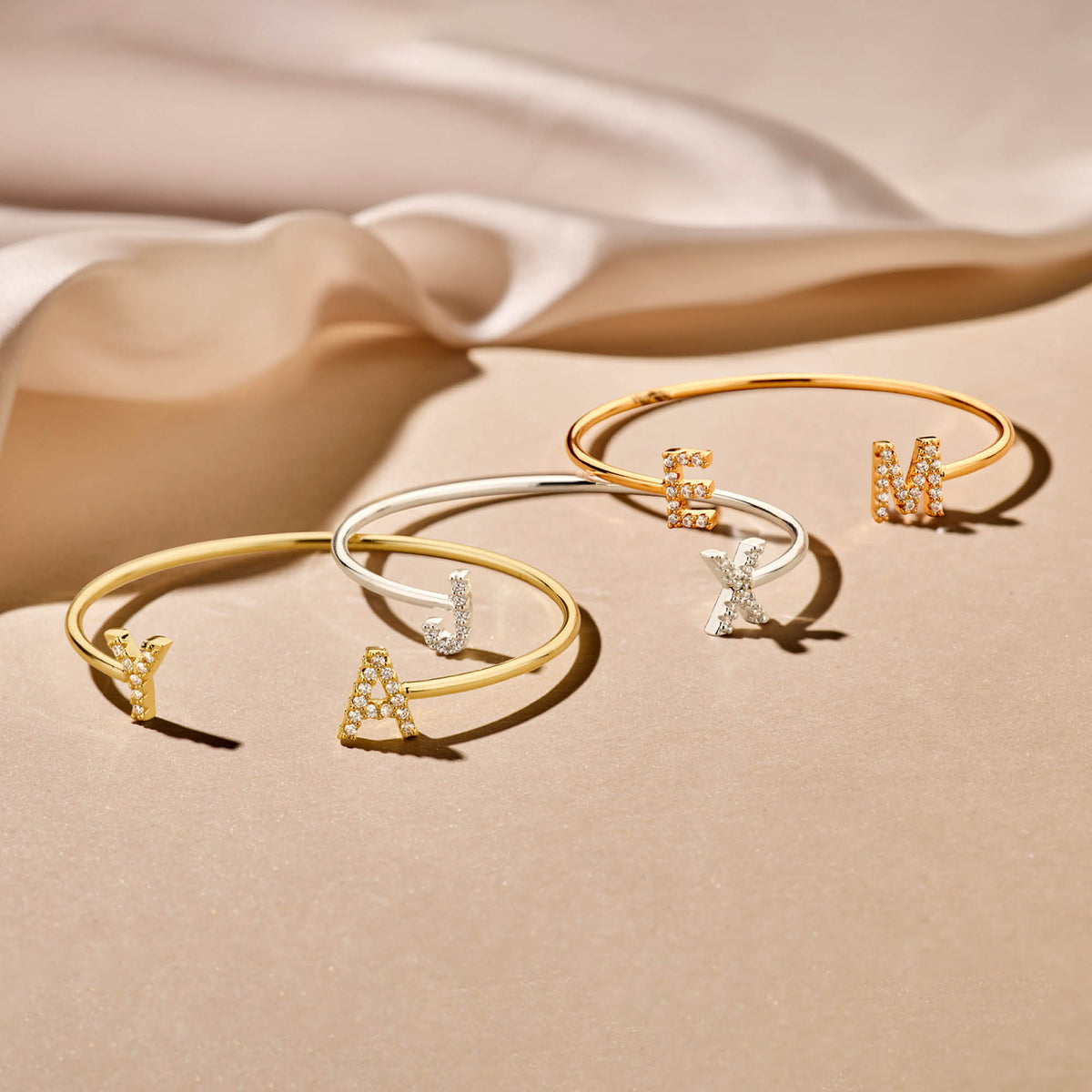 Drie open letterarmbanden in goudkleur, zilverkleur en rosékleur liggen op een beige ondergrond.