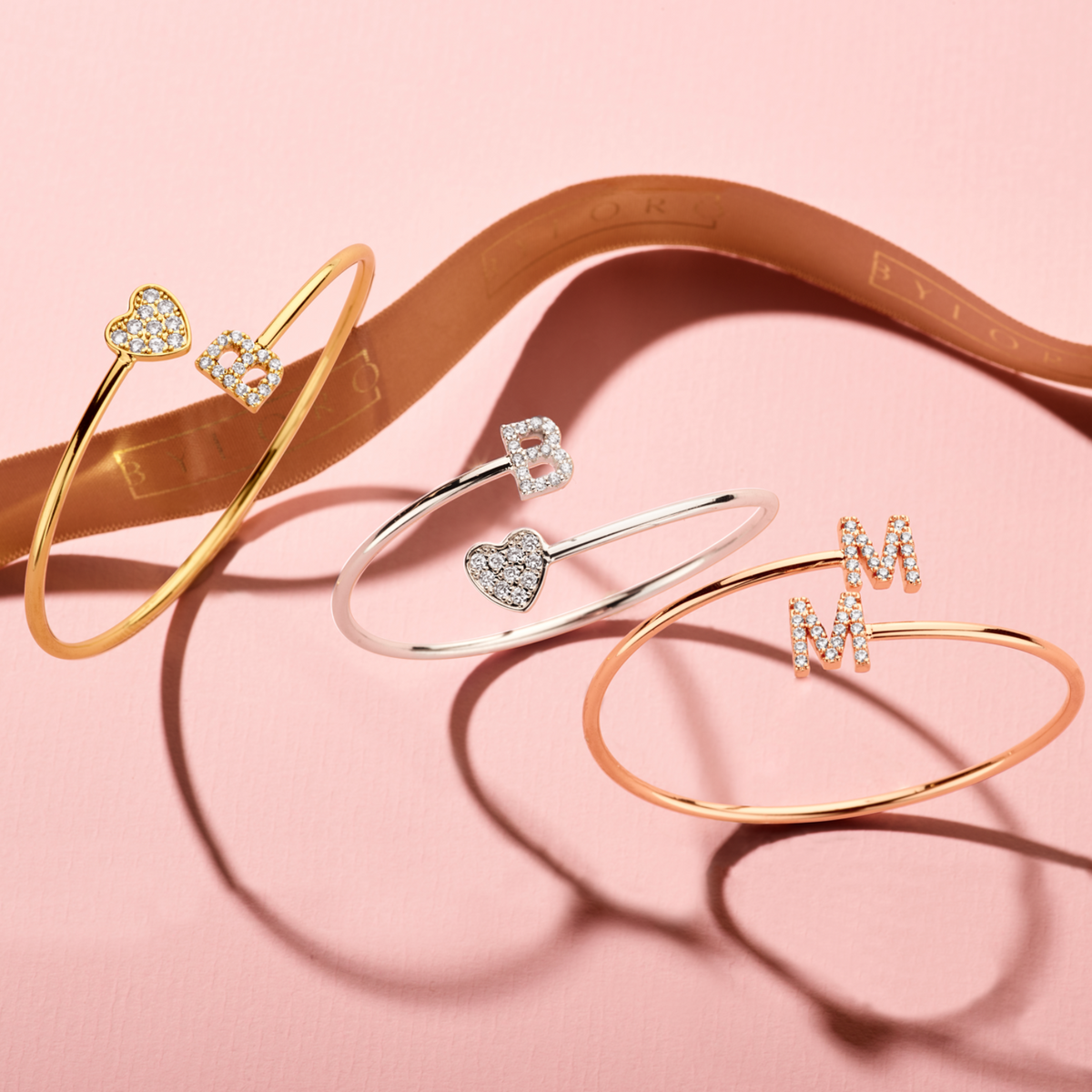 Drie open armbanden met glinsterende letters en hartjes liggen op roze stof naast een Byloro-lint.